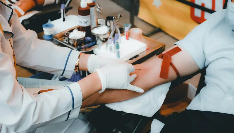 Ein Mitarbeiter des Gesundheitswesens, der Handschuhe trägt, bereitet sich darauf vor, einer Person, deren Arm mit einem Stauschlauch abgeschnürt ist, Blut abzunehmen; Probenröhrchen und weiteres Zubehör liegen auf einem Tablett in der Nähe bereit.
