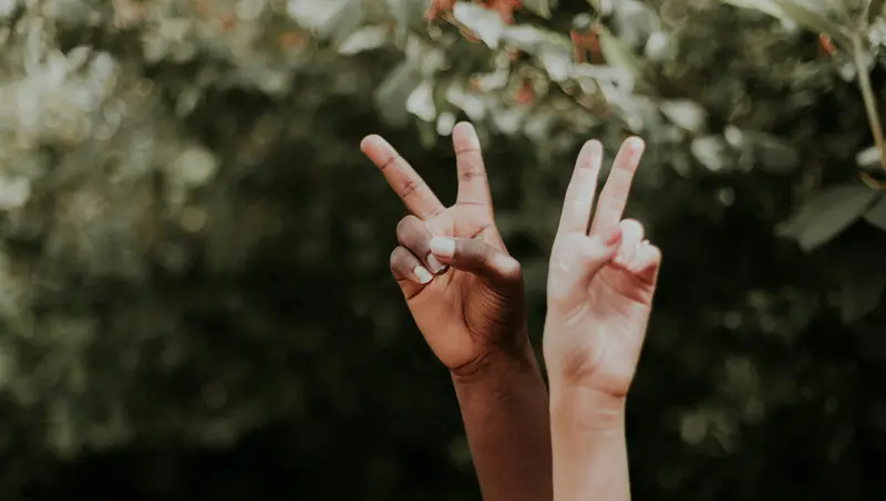 Duas mãos levantadas, de tons de pele diferentes, fazem o sinal da paz contra um fundo verde e folhoso difuso.