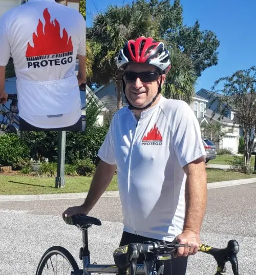 Ein Radfahrer mit rotem Helm steht mit seinem Rennrad da und trägt ein weißes PROTEGO-Trikot, auf dessen Rücken ein Einsatz mit dem großen PROTEGO-Flammenlogo zu sehen ist.