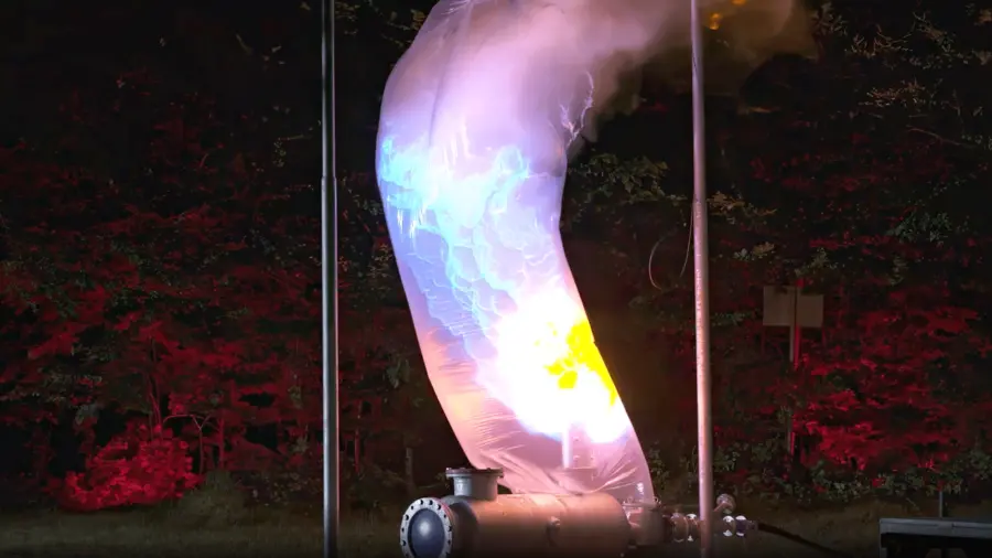 Nighttime industrial test showing a curved pipe releasing a large, brightly glowing flame or ignition plume inside a translucent protective sleeve, illuminated against a wooded background.
