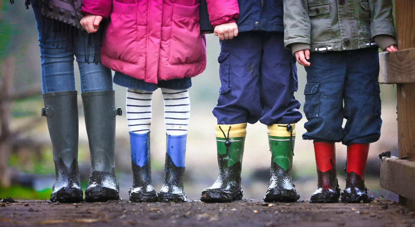 Nahaufnahme eines Erwachsenen und dreier Kinder, die in schlammigen Gummistiefeln stehen und von denen nur die Beine und Mäntel zu sehen sind.