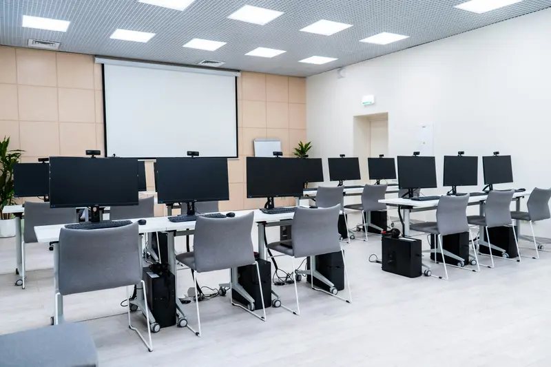 Une salle de informatique vide, avec des rangées de bureaux, des écrans d'ordinateur et un grand écran de projection à l'avant.