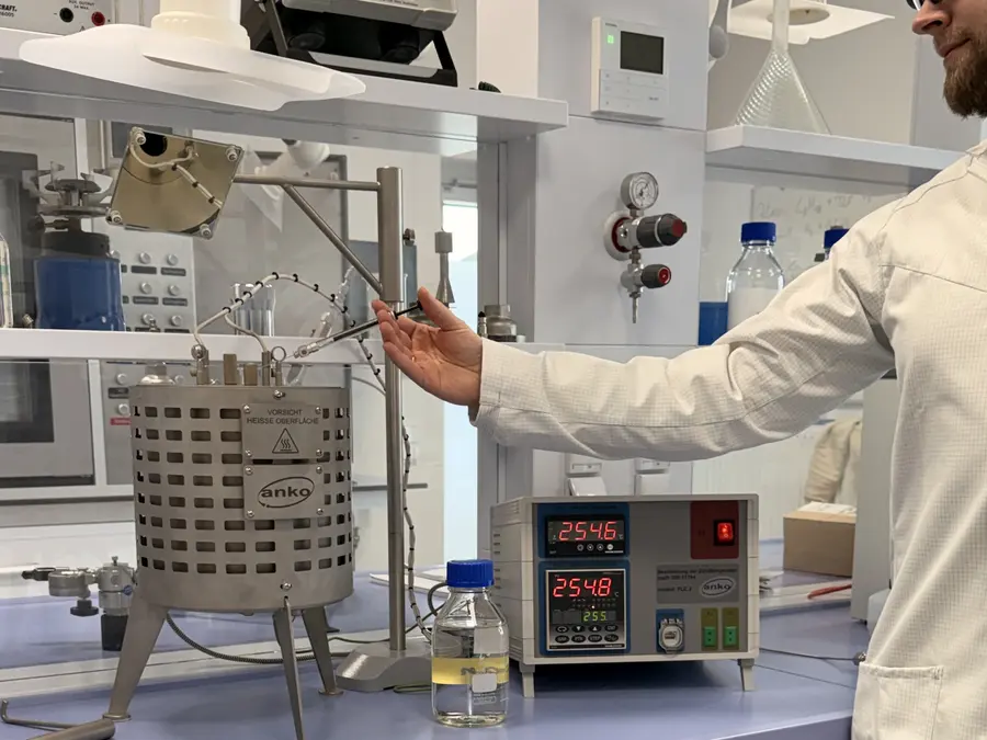 Una persona con bata de laboratorio maneja un depósito metálico calentado en un laboratorio, junto a una unidad de control digital y unos frascos sobre la mesa.