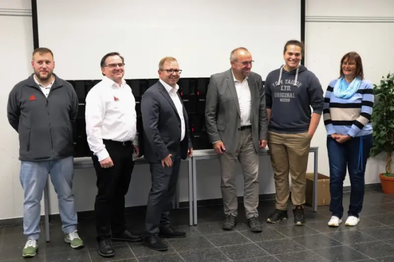 Six personnes posent en souriant pour une photo de groupe dans une salle de classe, devant une table sur laquelle sont alignés des mallettes noires contenant du matériel et un écran de projection.