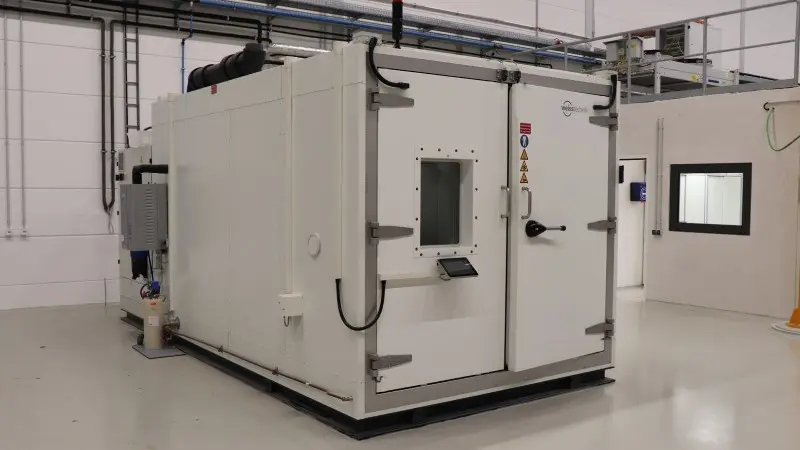 Large enclosed industrial test chamber with reinforced doors, viewing window, and warning labels installed inside a clean factory hall.