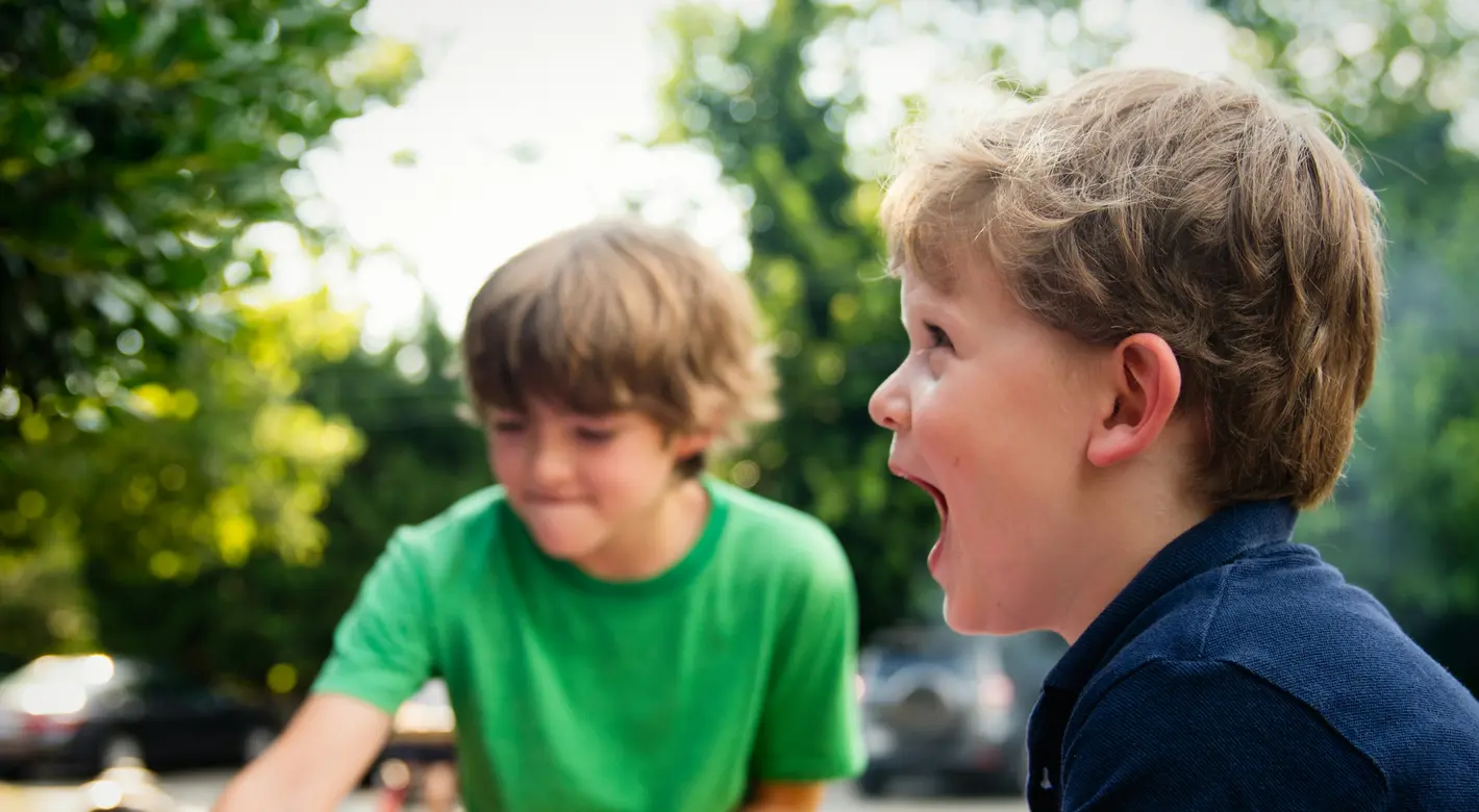 Zwei Jungen spielen draußen in einem kleinen rosa aufblasbaren Planschbecken; im Vordergrund lacht einer der Jungen.