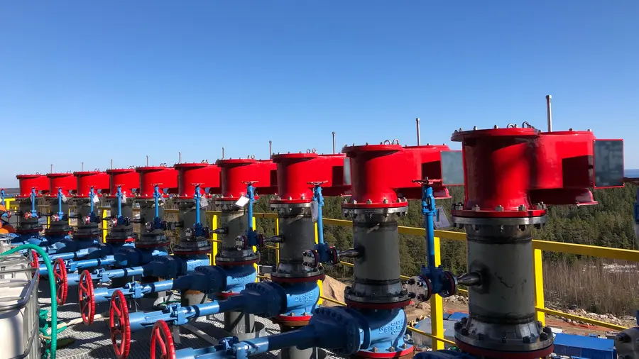Uma fileira de grandes válvulas de ventilação industriais vermelhas está disposta sobre tubulações azuis e volantes, em uma plataforma ao ar livre sob um céu azul límpido.