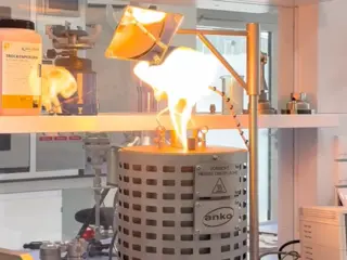 A laboratory burner flares with a bright yellow flame above a metal heating vessel under a mounted heat lamp.