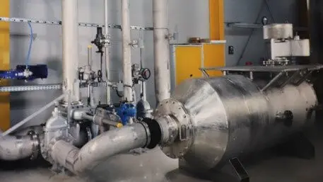 Industrial piping installation with a large cylindrical inline device connected to valves and instruments inside a processing or test facility.