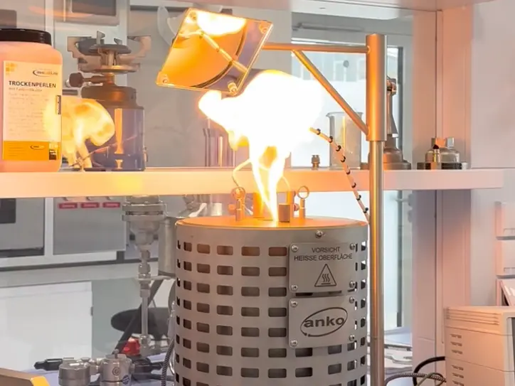 Un mechero de laboratorio arde con una llama de color amarillo brillante sobre un depósito metálico de calentamiento, bajo una lámpara de calor fijada al soporte.