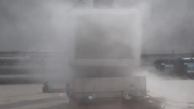 Outdoor industrial test setup with a large container largely obscured by a dense cloud of venting vapor or smoke during an active test.