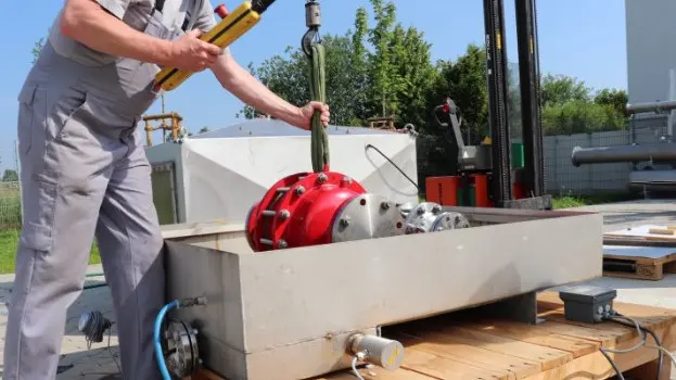 A technician lowers a large red industrial safety device into a rectangular outdoor test basin using a hoist, with cables and piping connected.