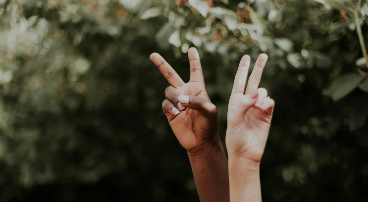 Zwei erhobene Hände unterschiedlicher Hautfarbe formen Peace-Zeichen vor einem verschwommenen, grünen, blättrigen Hintergrund.
