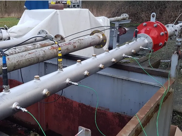 Montaje de ensayo industrial al aire libre con un tubo horizontal sobre un foso de contención abierto, que incluye un equipo con brida roja y varios sensores conectados mediante cables verdes.