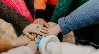 Several people stack their hands together in a team huddle, with colorful sweaters visible around the circle.