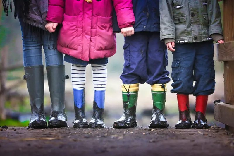 Primer plano de un adulto y tres niños con botas de agua embarradas, en el que solo se ven sus piernas y sus abrigos.