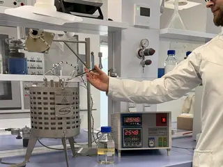 A person in a lab coat operates a heated metal vessel in a laboratory, with a digital control unit and bottles on the bench.