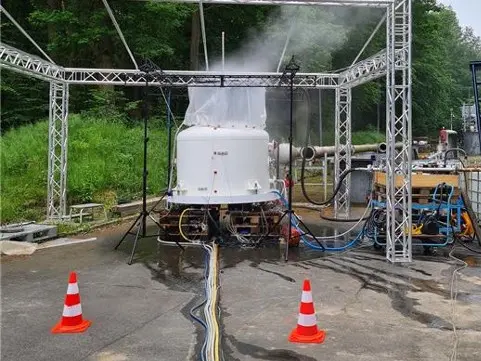Instalación de pruebas industriales al aire libre con un depósito blanco del que sale vapor bajo una estructura de vigas metálicas, rodeada de mangueras, equipos y conos de seguridad.