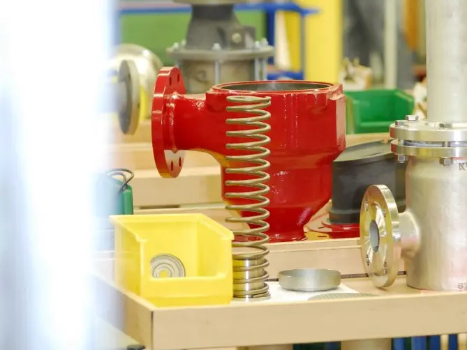A red industrial valve housing with a large metal spring and various mechanical parts laid out on a workbench.