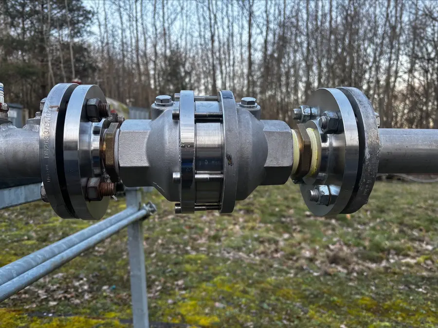 Close-up of a metal pipe coupling with flanges and bolts, installed outdoors with trees in the background.