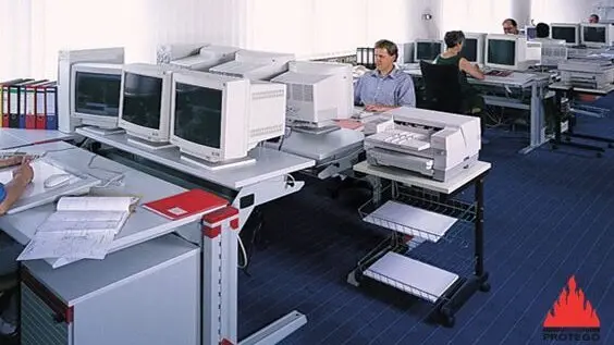 A small open-plan office with several people working at desks surrounded by multiple desktop computers and paperwork. 