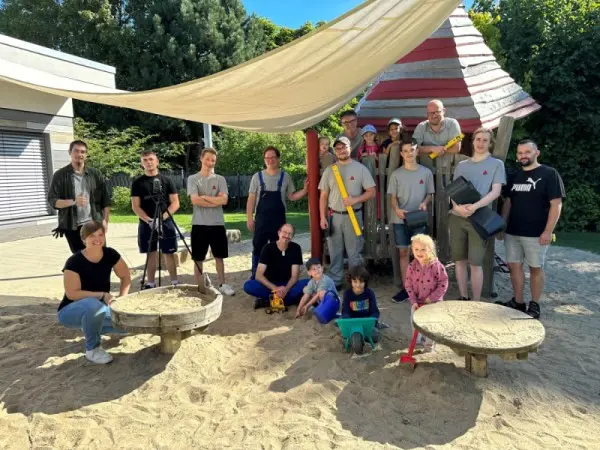 A group of adults and children pose together in a sunny playground sand area under a large shade sail, with play equipment behind them.