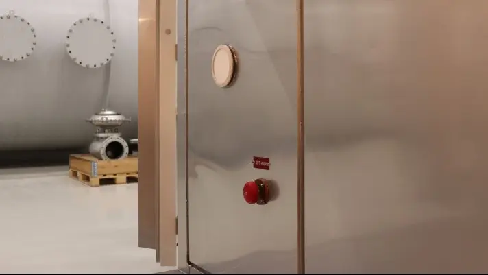Stainless steel industrial enclosure door with an emergency stop button in the foreground, with a large pressure vessel visible inside a factory hall.