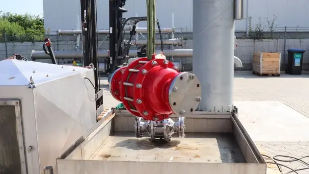 A large red industrial inline safety device is suspended by lifting straps above a metal test basin at an outdoor facility, with piping and equipment nearby.