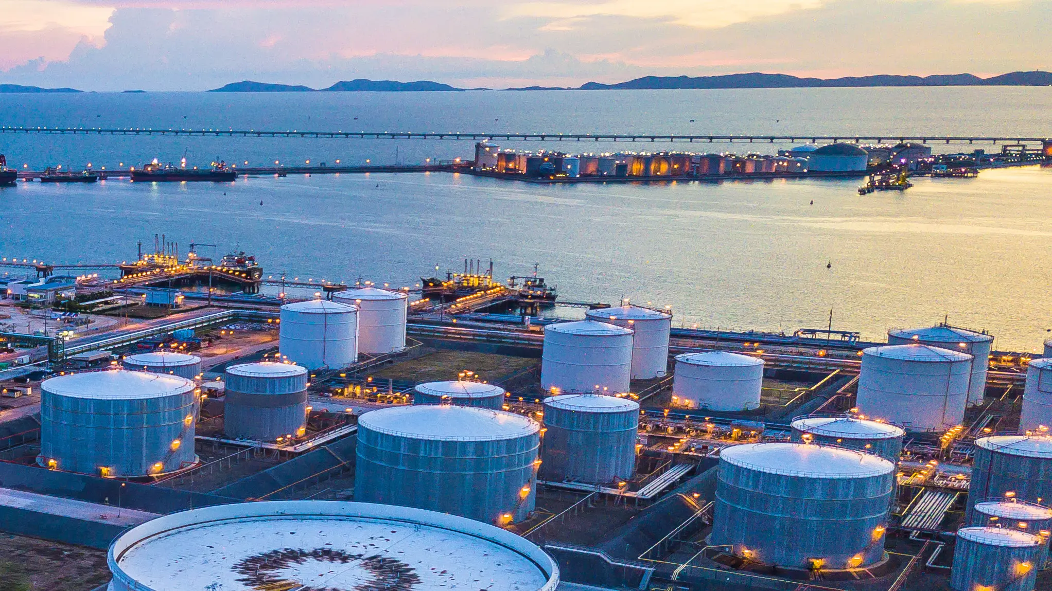 An oil and gas industry complex with large storage tanks near a harbor at dusk