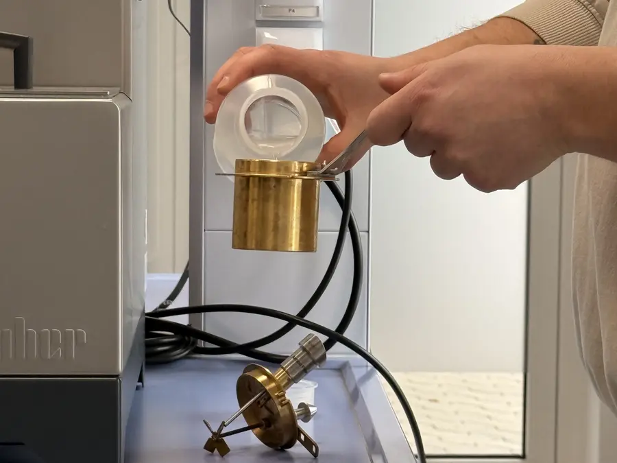 A person holds a small brass cup with tongs while pouring a clear liquid from a plastic container in a lab.