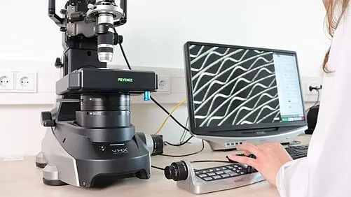 A person operates a microscope imaging system beside a monitor showing a magnified pattern of crisscrossing lines.