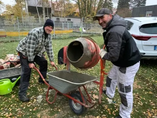 Two workers smile outdoors beside a wheelbarrow and a cement mixer, surrounded by fallen leaves and a parked car.