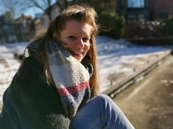 A smiling woman in a thick striped scarf sits outdoors on a sunny winter day, with snow and a walkway blurred in the background.