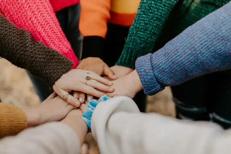 Several people stack their hands together in a team huddle, with colorful sweaters visible around the circle.