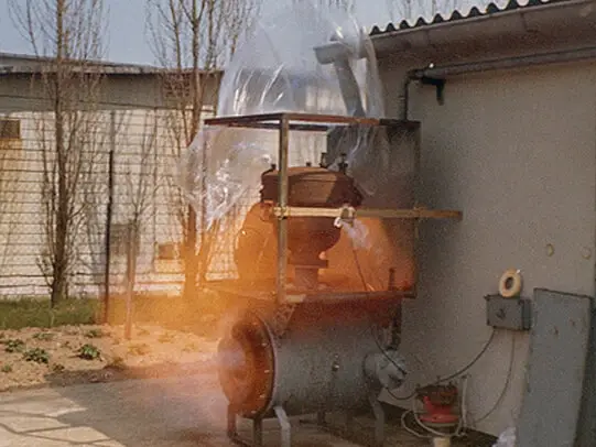 An outdoor industrial test setup with a cylindrical tank and piping is partially enclosed in a clear plastic sheet; flames and smoke are visible at the base, indicating a controlled fire or explosion test.