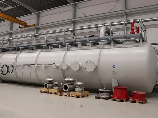 Large horizontal industrial pressure vessel inside a factory hall, equipped with multiple flanged nozzles, valves on pallets, and an elevated service platform.