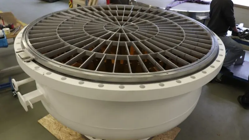 A large circular industrial component with a radial metal grate sits on a workshop floor, with a worker in the background.