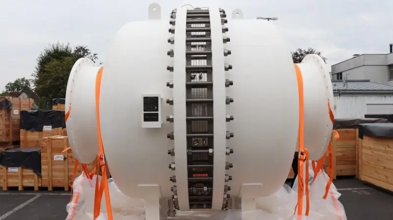 A large white cylindrical industrial vessel sits outdoors, secured with orange straps among shipping crates.