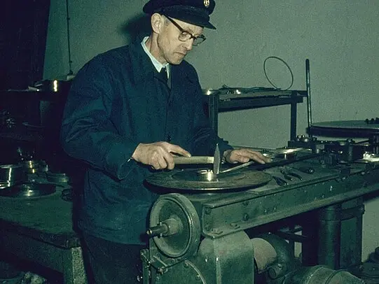A technician in a workshop carefully measures a circular metal component on a workbench using hand tools and gauges.