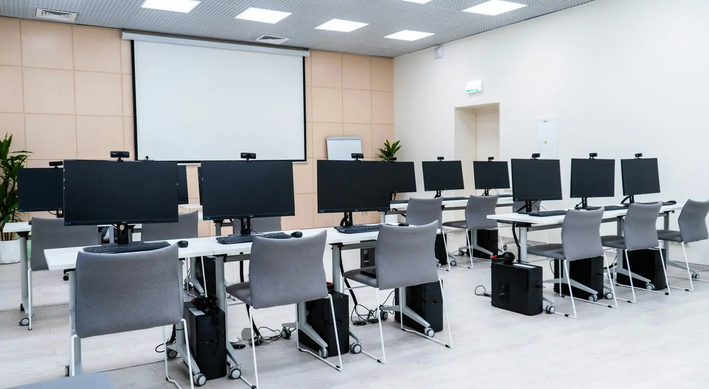 An empty computer lab classroom with rows of desks, desktop monitors, and a large projector screen at the front.