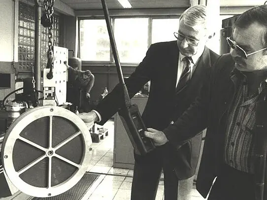 Two men in suits examine a large circular industrial component in a workshop, one holding a handheld measuring device while a worker stands in the background.