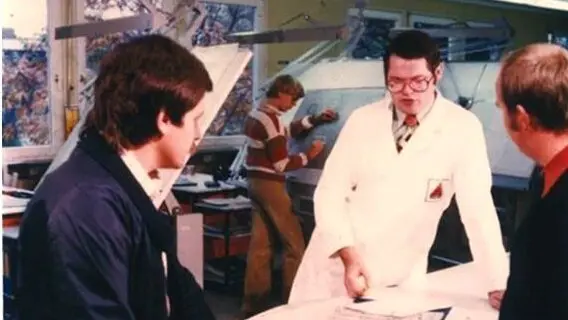 A man in a white lab coat discusses a document with two colleagues in an office, while another person works at a drafting table in the background.